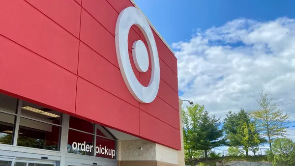 A red storefront entrance with a white dot-in-circle logo and an "order pickup" sign on the door. Trees and blue sky with white clouds on the right.