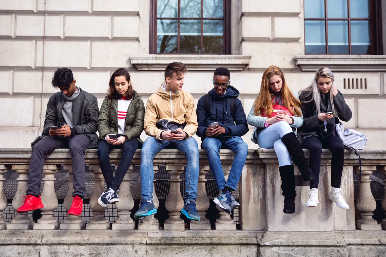 A row of six teenagers are sitting on a outdoor bench. Most are looking down at cellphones in their hands.