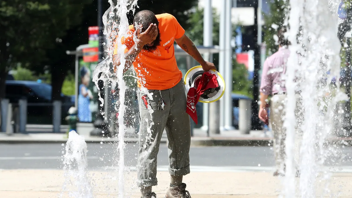 A construction worker stands near a fountain spraying water vertically, splashing it onto their face