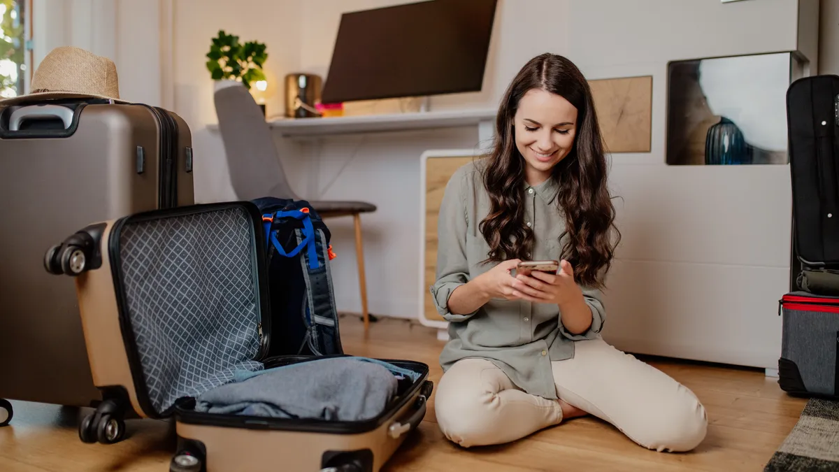 Young smiling person using mobile phone at home while packing a suitcase.