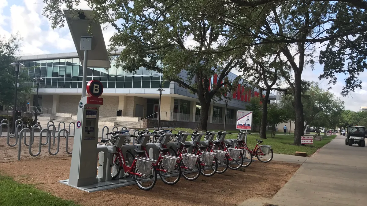 Eight bicycles docked at a station under trees next to a sidewalk.