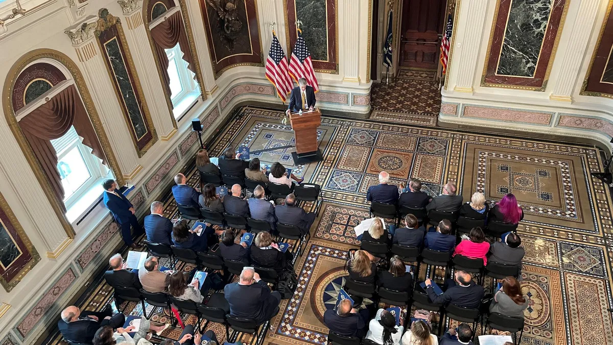 A person standing at a podium speaking before a group of people in a room with an ornate floor.