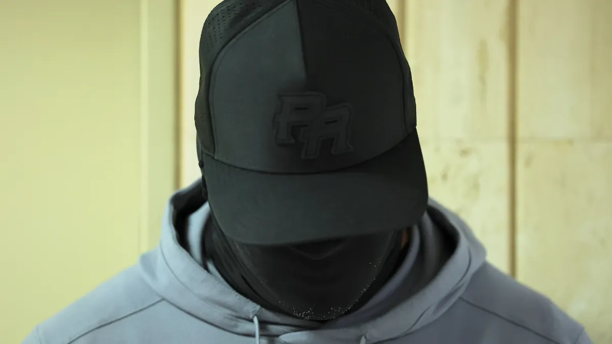 Close-up of a person's head covered with black mask, wearing black baseball hat and gray hoodie.