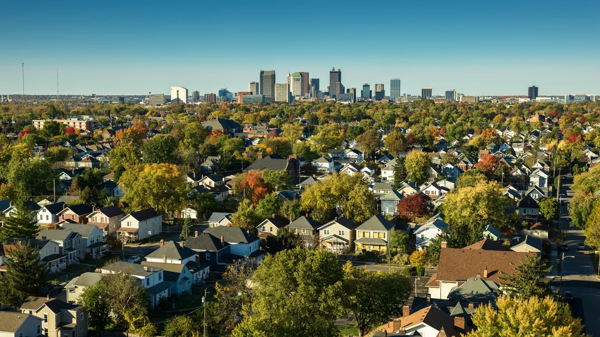 Aerial shot of homes and trees. In the distance, high-rises form a city skyline against clear blue skies.