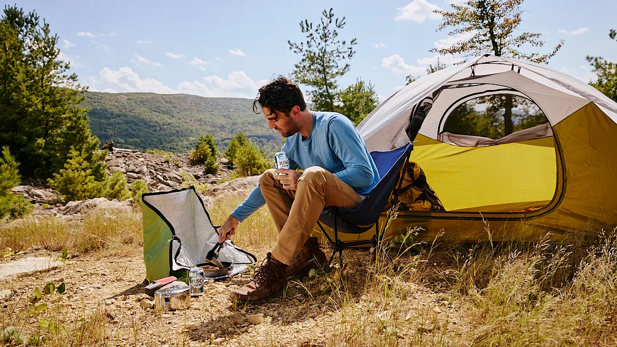 A person cooks a meal using a flameless cooker while holding a can of Busch Light outdoors.