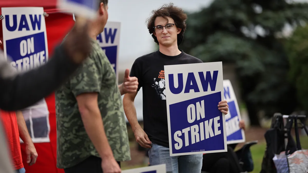 Two autoworkers look at each other with "UAW on Strike" signs in their hands and plastered on makeshift tents.