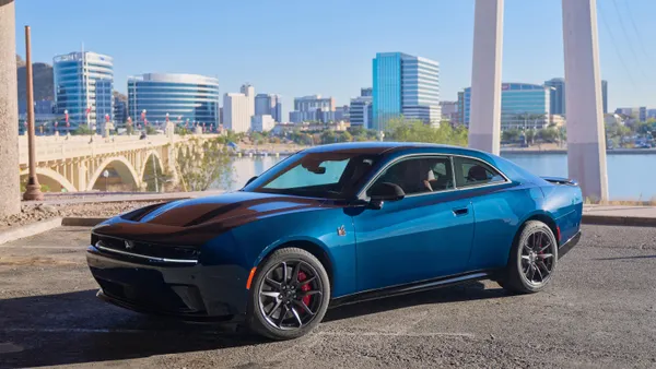 A 2-door blue 2026 Dodge Charger Daytona Scat Pack electric sports car parked outdoors with a city skyline and bridge in the background.