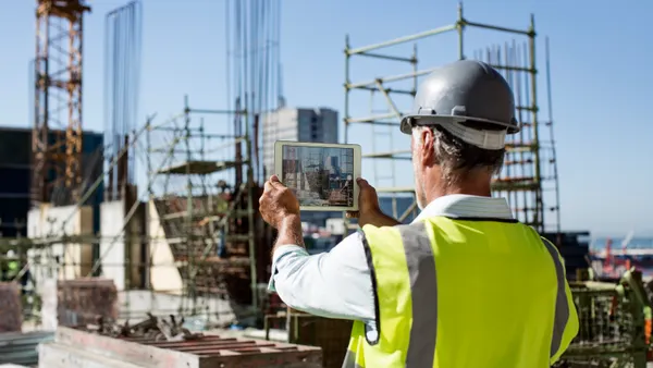 A person in safety gear, which includes a reflective vest, hardhat and safety glasses, holds a tablet up to take a photo of a construction site.