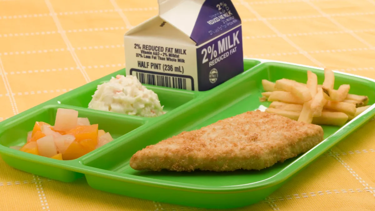 A green cafeteria tray rests on a yellow tablecloth. On the tray are French fries, a small carton of 2% milk, fruit slices and coleslaw.