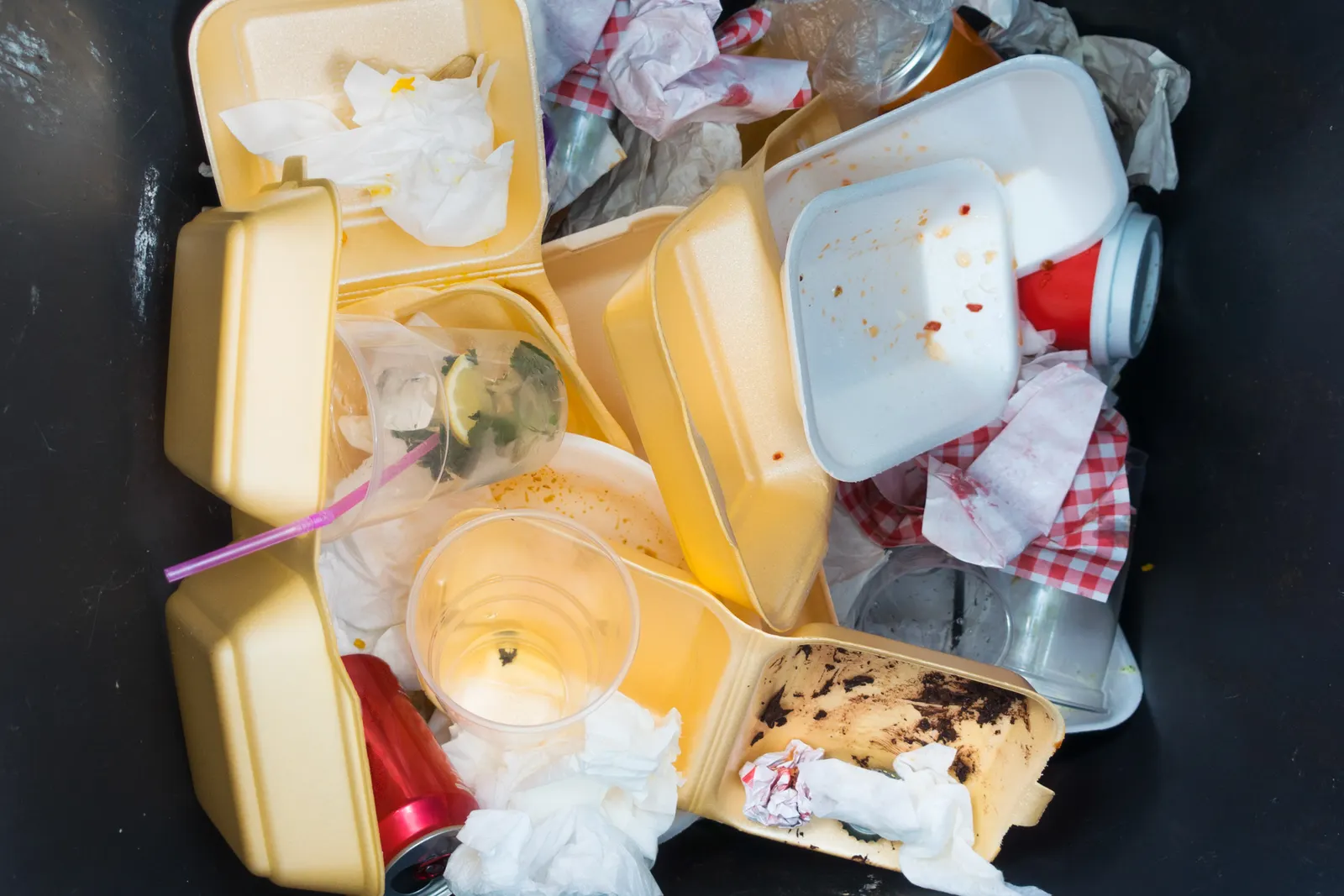 An overlooking angle into a black trash can filled with yellow styrofoam fast food containers, wrappers, and soda cans.