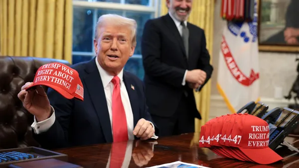 Donald Trump holds up a hat as he signs an executive order while sitting at his desk in the Oval Office