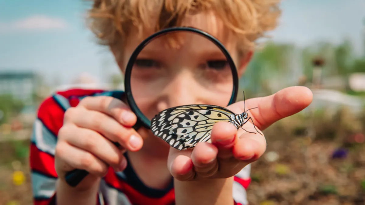 A student is holding a butterfly outside. The student has a magnifying glass to their face.