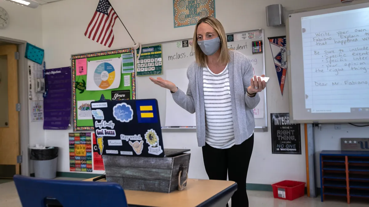 An adult stands in front of an open laptop on a table in a classroom. The adult is wearing a face mask