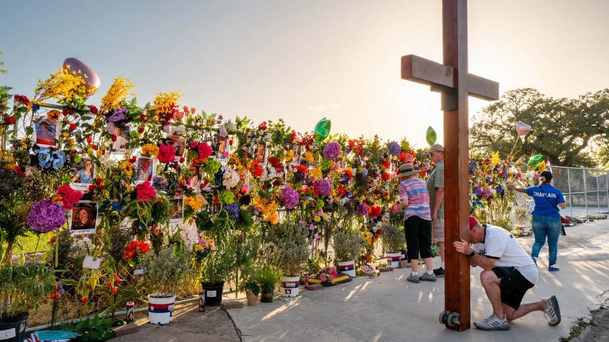 A person kneels at a large wooden cross as two people look at a fence covered with flowers and photos.