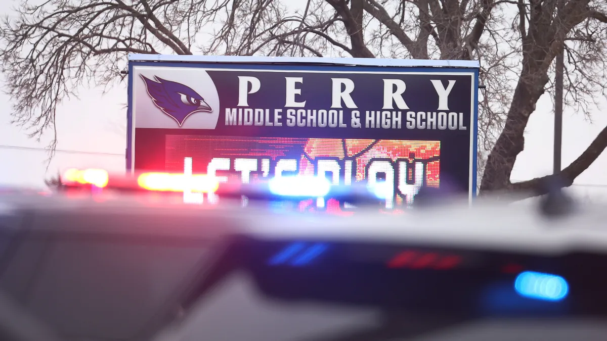 Out-of-focus police lights are seen in front of a sign for Perry Middle School and High School stands against an overcast winter sky and leafless trees.