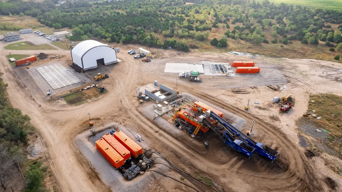 Aerial view of an industrial facility with a scraped earth ground and large shed. Conveyor belts also load material into one gravel-lined spot.