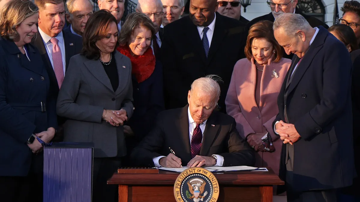 President Joe Biden is pictured seated with various legislators standing around him as he signs a document.