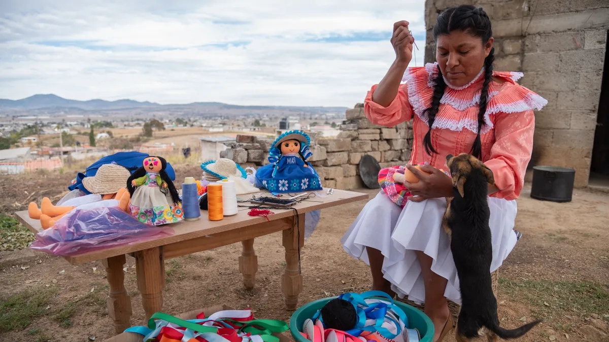 A person is seen sewing a stuffed doll outdoors, next to buckets of fabric. A dog is with them.