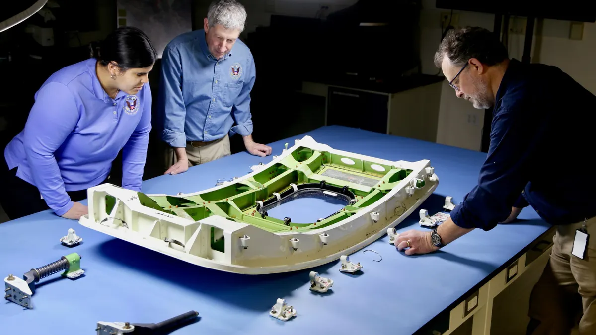 Three people assessing a display of a plane's doorplug and its parts on a blue table.