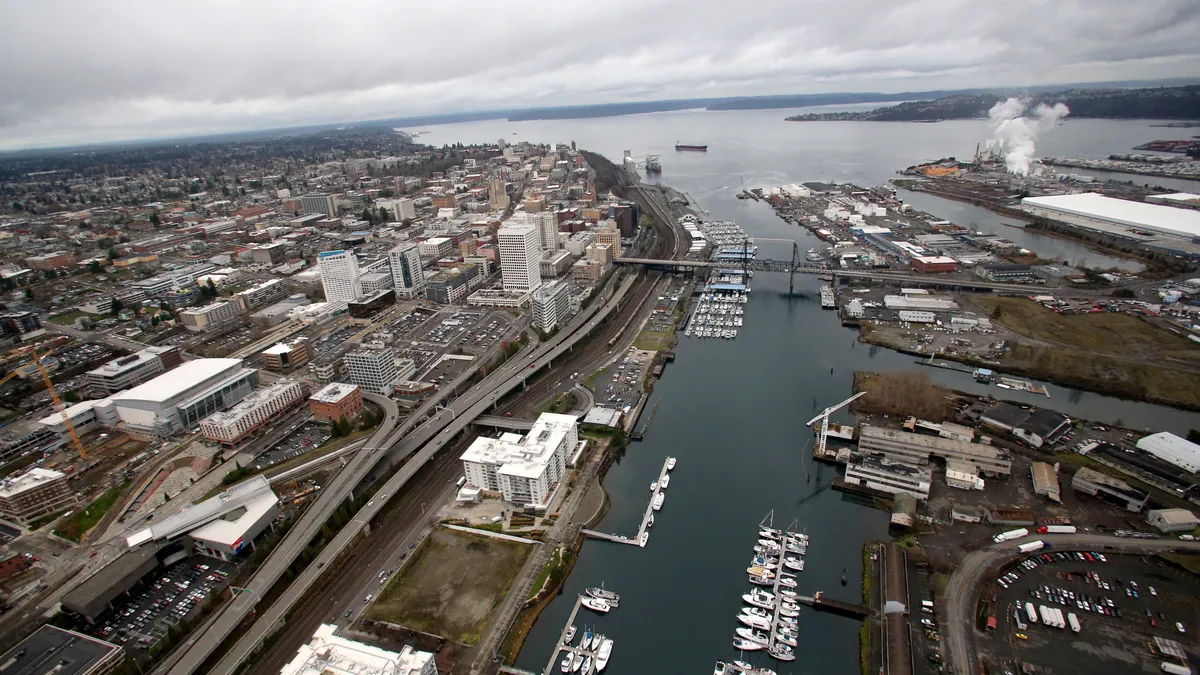 Aerial photo shows a channel filled with boats leading out to a large body of water, with buildings and roads on either side.