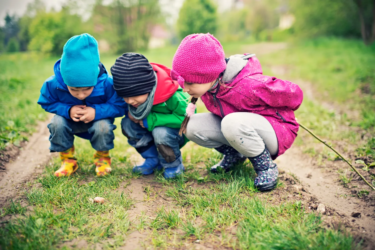 Three young students are outside and leaning close to ground. They are looking down at the grass.