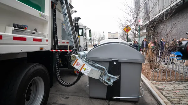 A mechanical arm on a garbage truck grabs a large waste bin sitting on the street near a sidewalk.