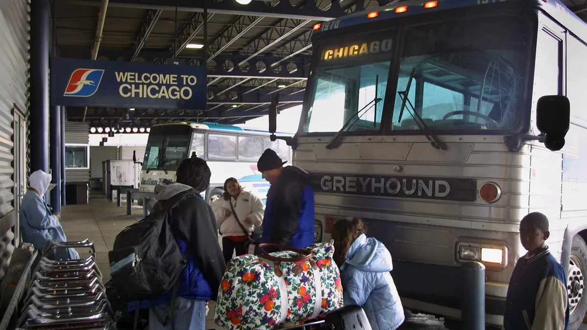 Passengers  with luggage standing by buses under a shed.