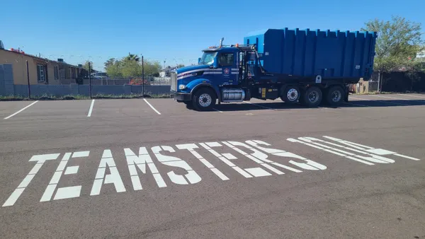 A waste truck sits in a parking lot with the words 'TEAMSTERS 104' painted on the pavement in front of it.