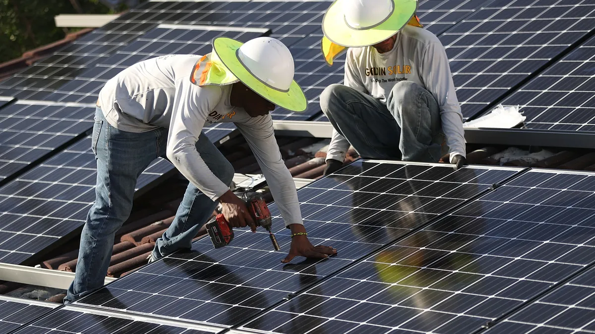 Two people install rooftop solar on a building. One person is holding the panel and the other person is holding a drill.