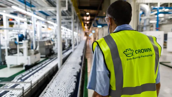 A person wearing a bright yellow vest with Crown branding on the back stands near a can manufacturing line.