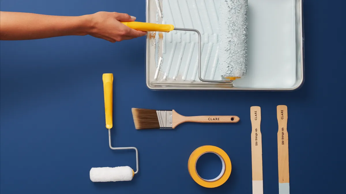 A hand holds a yellow paint roller on top of a tray with light blue paint