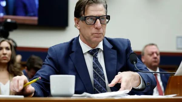 A person wearing a suit and tie holds a pencil and shuffles papers while sitting at a desk with two people sitting behind him.