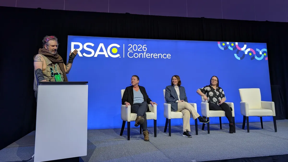 Three women sit on a stage and one man stands on the stage with them. Behind them, a blue backdrop displays a logo reading "RSAC 2026 Conference."