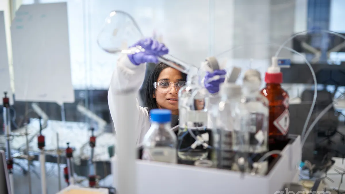 Image of a person wearing lab goggles and pouring a liquid out of a beaker.