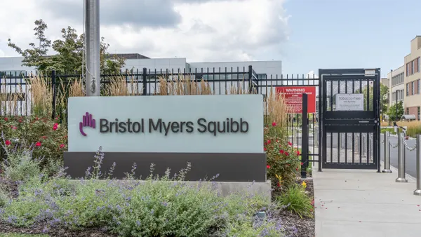 A white building entrance sign reads "Bristol Myers Squibb," with a barbed fence behind it surrounded by flowers.