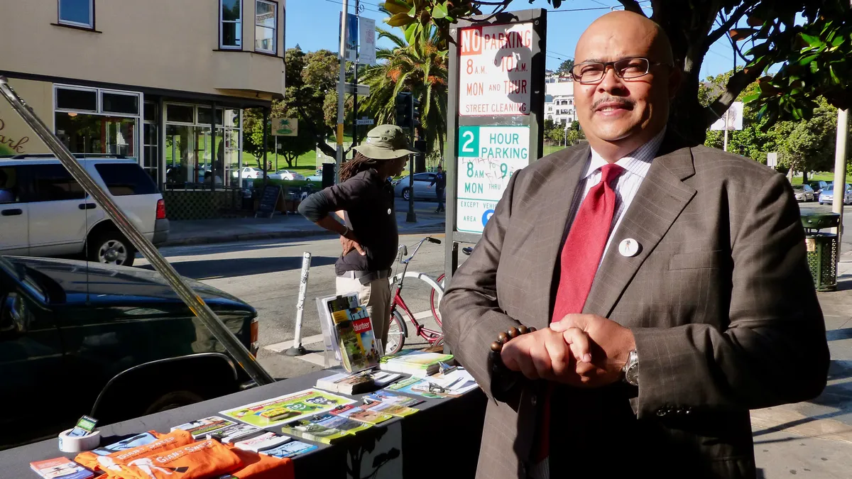 Man in suit, with a mustache, standing in front of a table with pamphlets under a tree on a city sidewalk