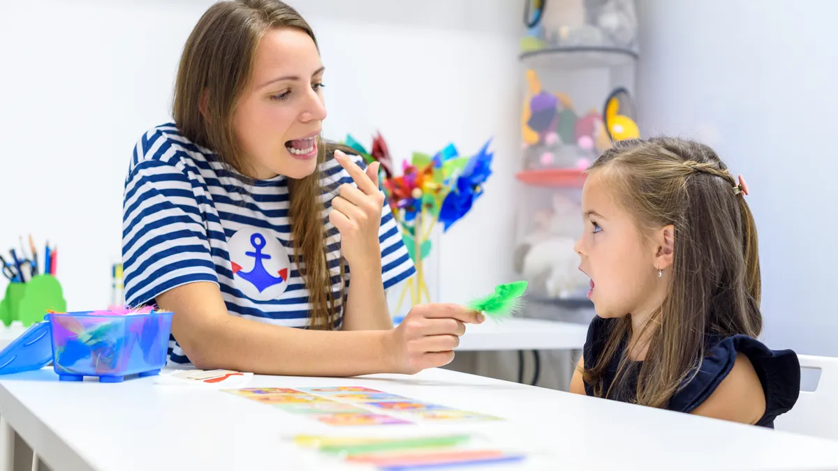 An adult and young student sit at a desk. They are looking at each other and the adult is pointing to their mouth and has their mouth open.