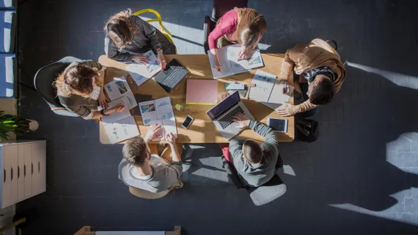 Overhead view of six students seated around a rectangular wooden table, working with open books, papers, and laptops. The scene is brightly lit with natural light from the side. Items like a smartphone, notebook, pens, and folders are scattered on the table. 