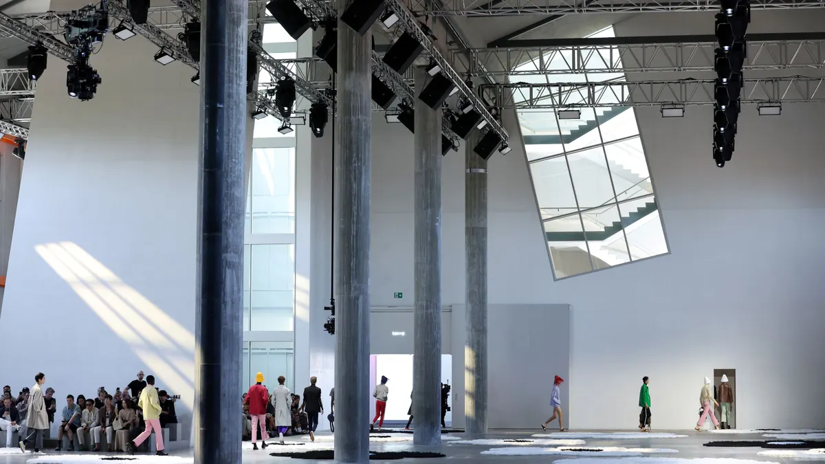 A line of models walk during the day in a cavernous room flanked by tall pillars.
