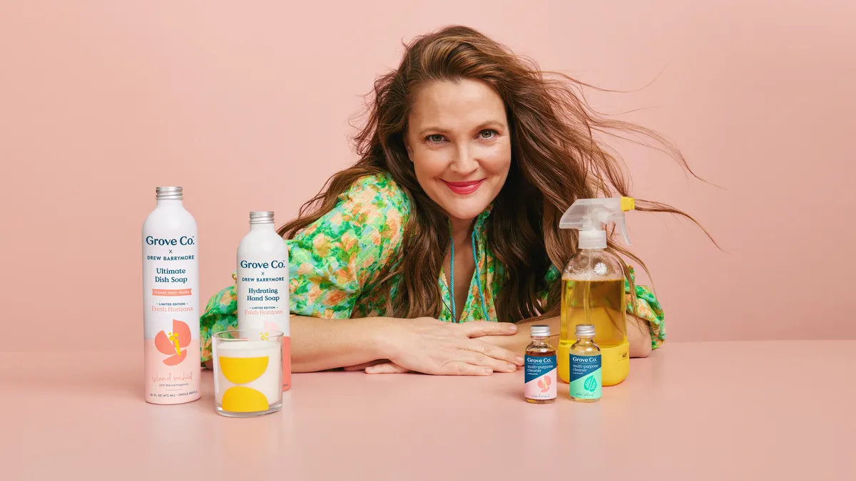 Drew Barrymore, with light brown wavy hair, smiles at a camera surrounded by cleaning bottles and a pink background.