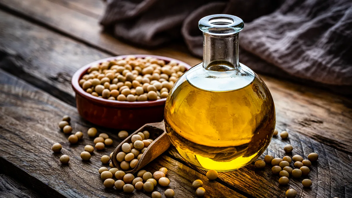 A close up view of a glass bottle filled with soy oil on a rustic wooden table with a bowl with dried soybeans on its left.