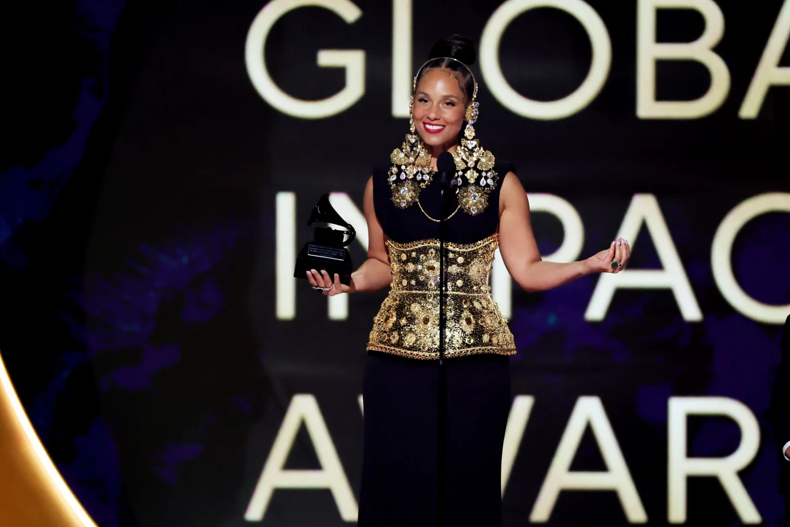 A person in a gold jeweled headdress, gold waist trainer and black dress stands on a stage holding an award.