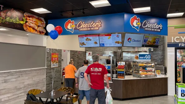 A photo of several people standing in line at a counter inside a store. A sign over the counter says Chester's Chicken on the Run.
