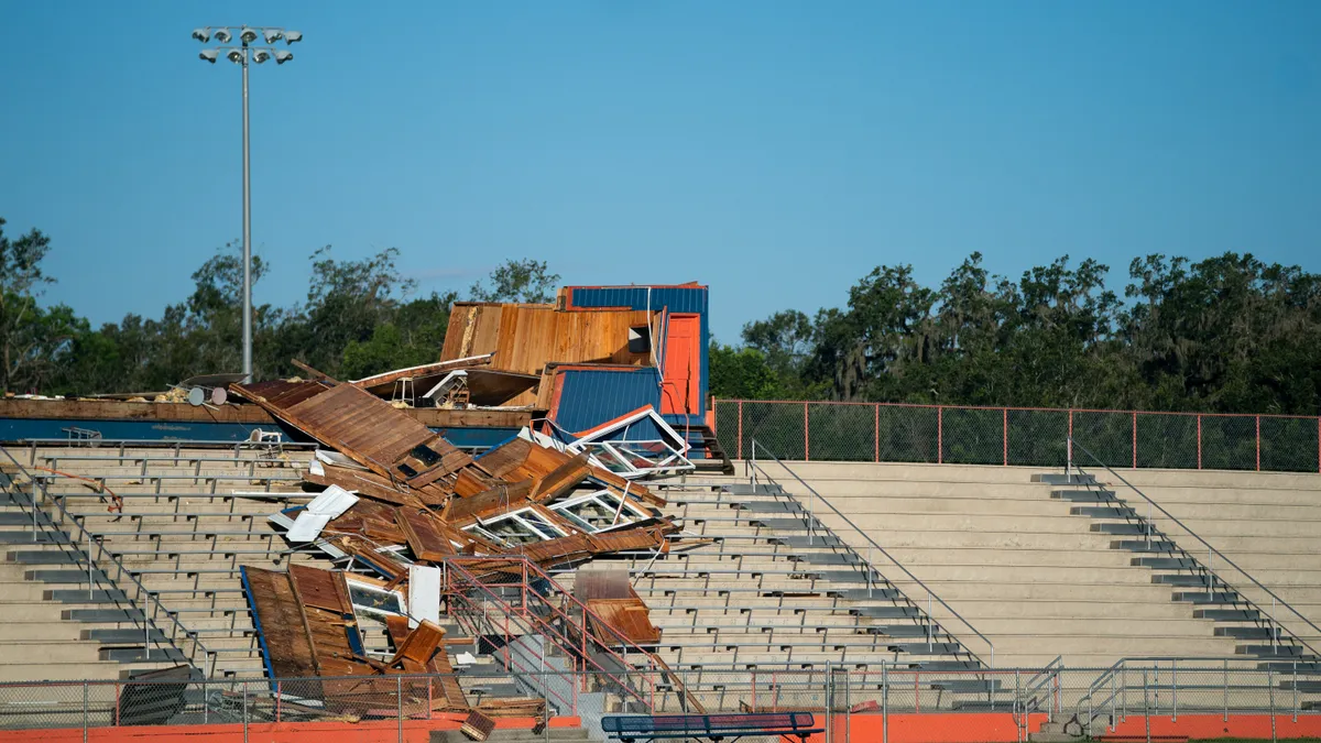 An outdoor  high school football stadium is shown with a wooden press box demolished at the top of the stadium