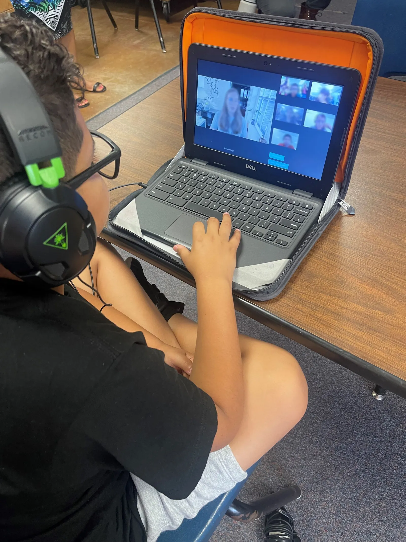 A young student wears headphones while working on a laptop during an online tutoring session. The laptop screen shows a tutor talking with other students on the same platform.