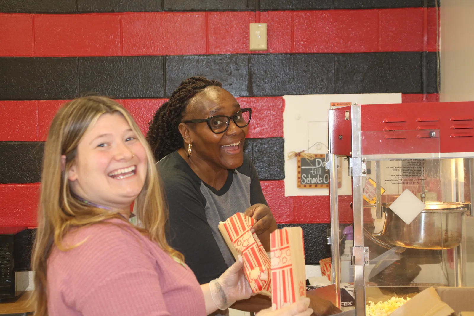 Two woman school staffers prepare popcorn for a party thrown in honor of students. They are scooping popcorn from a popping machine into bags, against the backdrop of a red and black brick wall.