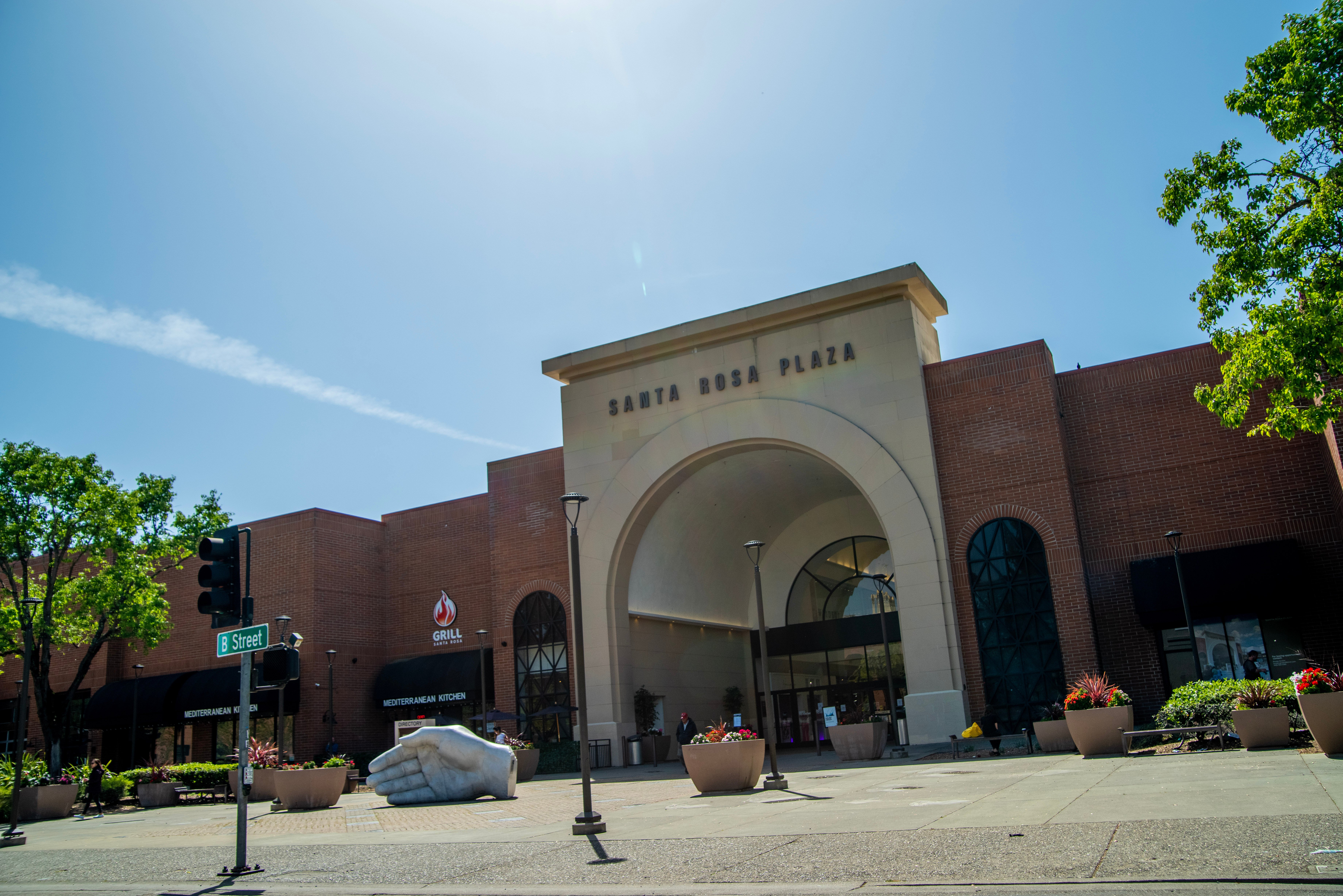 The arched entrance to a building reads "Santa Rosa Plaza," with trees, potted flowers and a sculpture of a hand in the foreground, and cloud-streaked blue sky in the background.