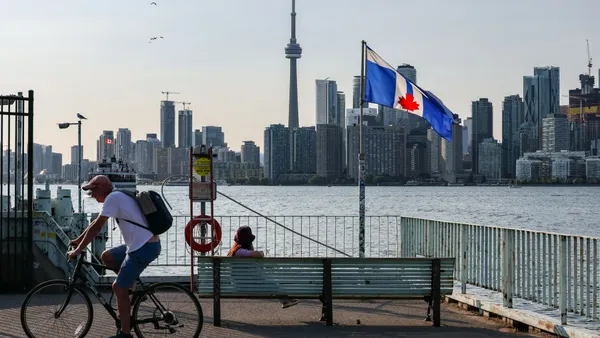 One person sits on a bicycle and another on a bench near a waterfront spot with the Toronto skyline in the distance.