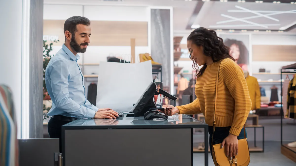 Young Woman at Counter Buys Clothes from Friendly Retail Sales Assistant, Paying with Contactless NFC Smartphone Touching Terminal.