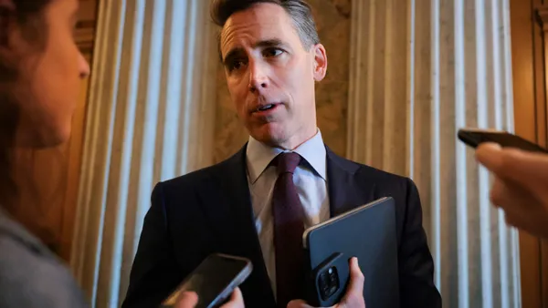 A man in a suit talks to reporters holding their phones close to him.